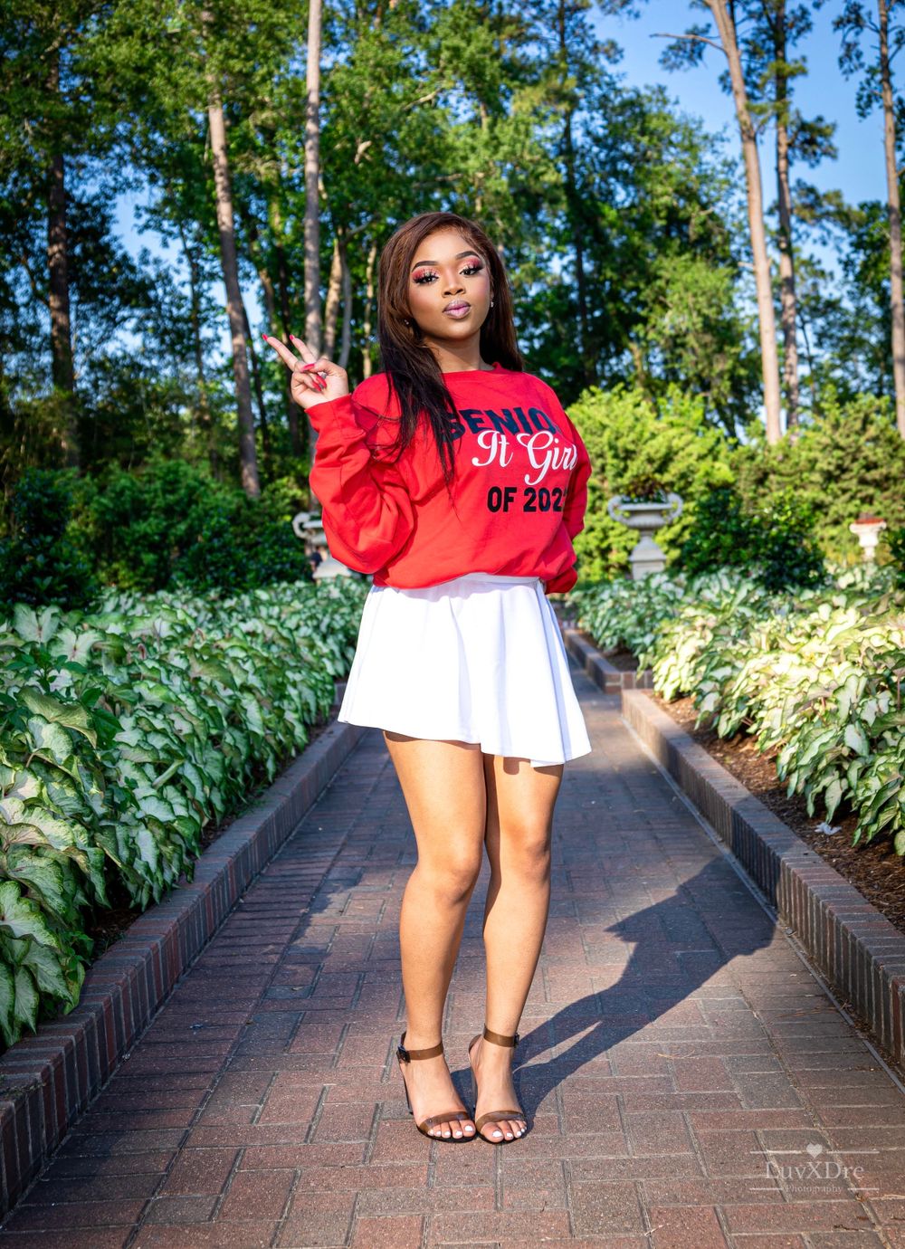 Graduate in red Senior sweatshirt and white skirt posing on garden pathway.
