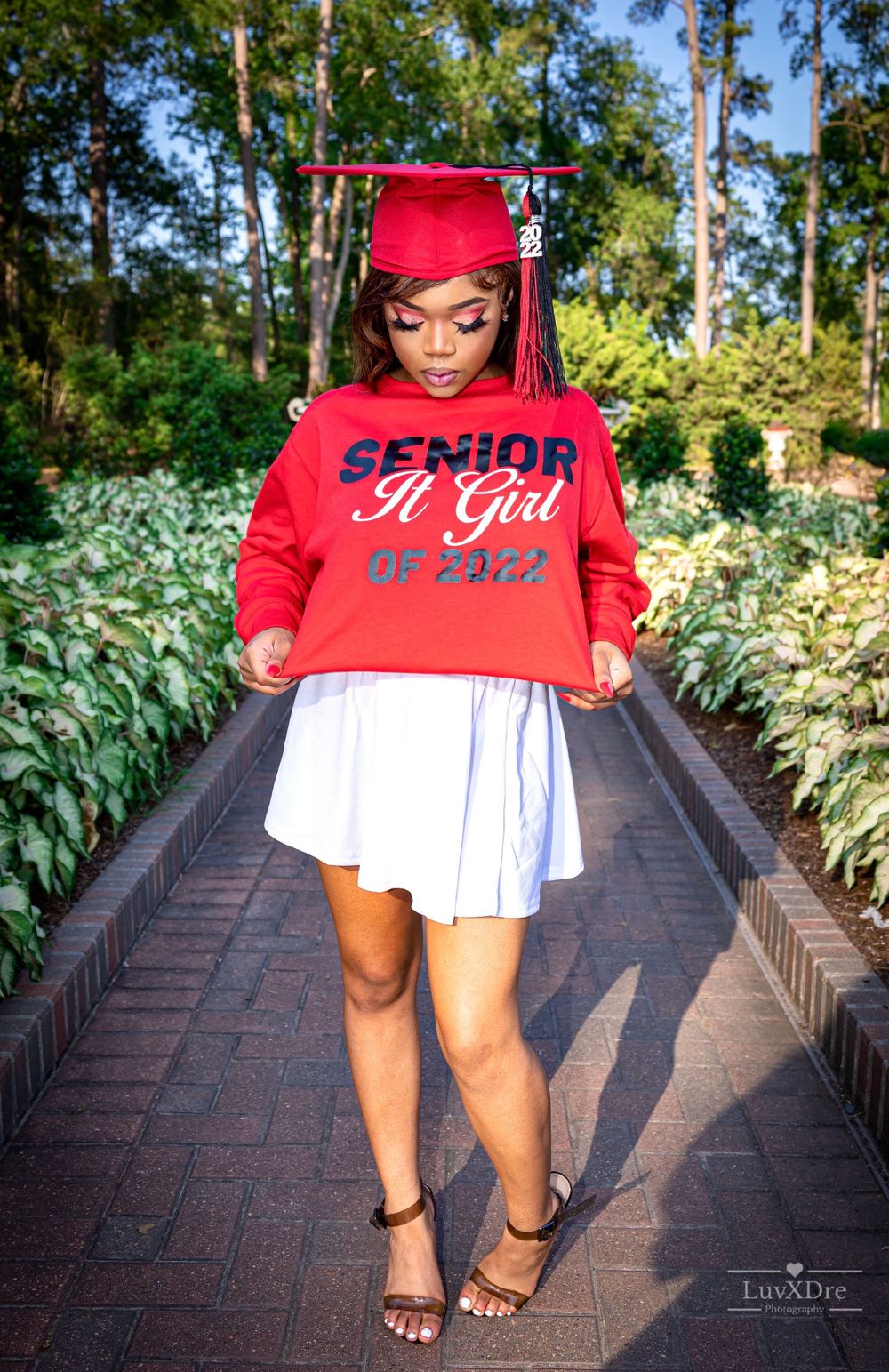 Graduate in red cap and sweatshirt posing on brick pathway surrounded by greenery.