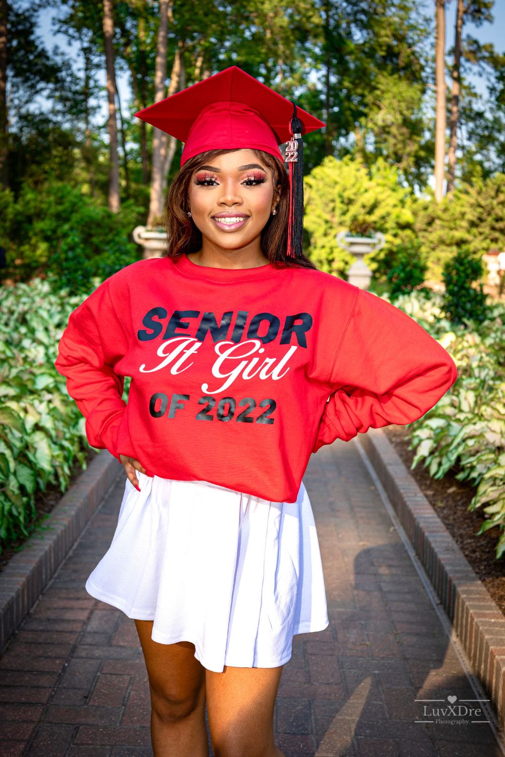 Graduate in red cap and sweatshirt with white skirt smiling on brick path.