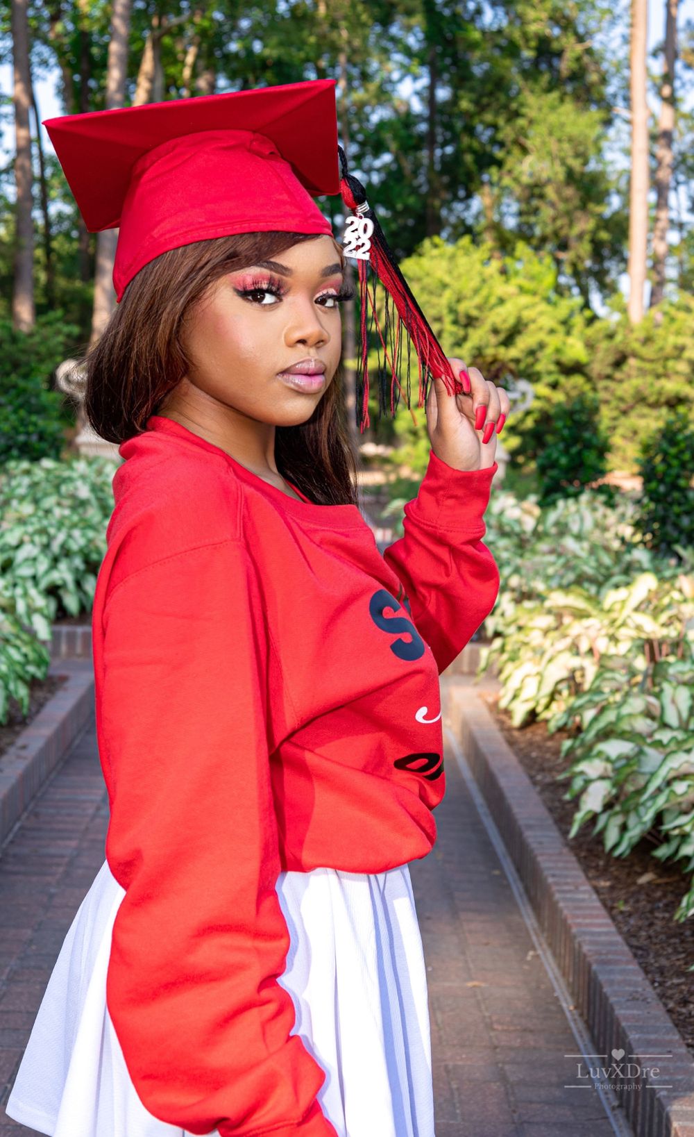 Graduate in red cap and sweatshirt poses against garden backdrop.