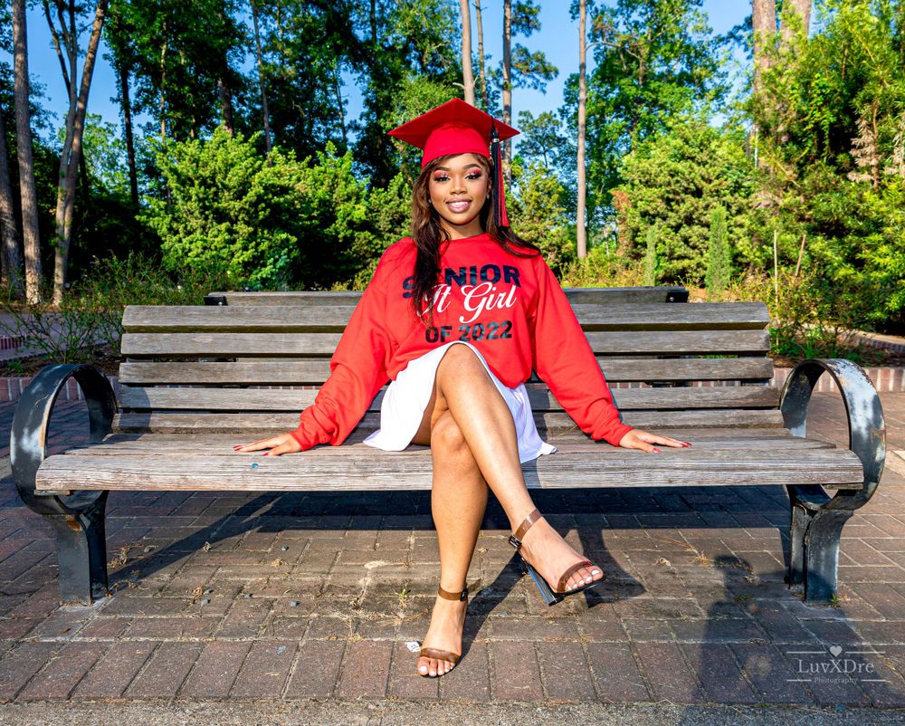 Graduate sitting on park bench in red sweatshirt and white skirt with heels.