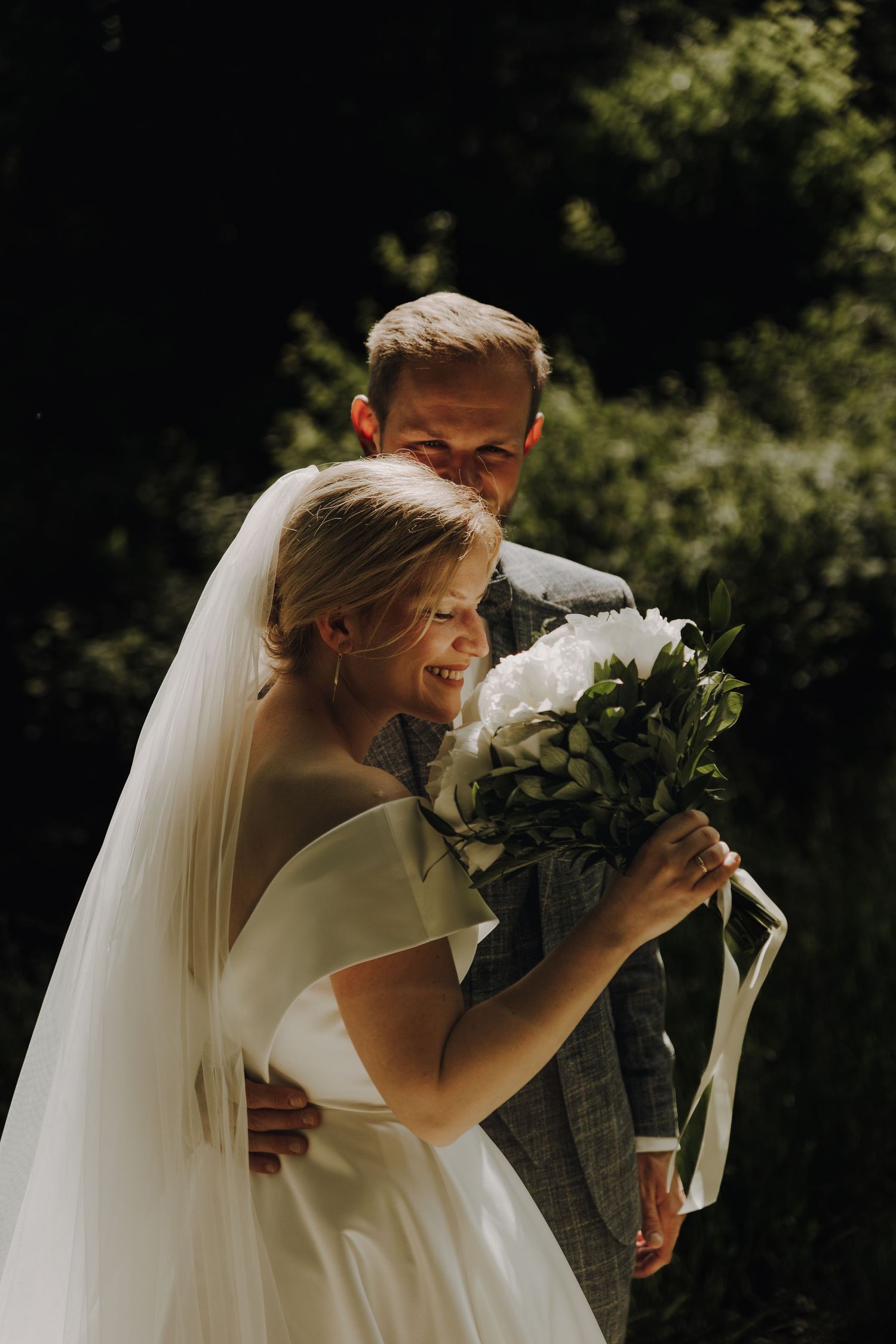 Sunlit bride holding white bouquet while sharing an intimate moment outdoors.