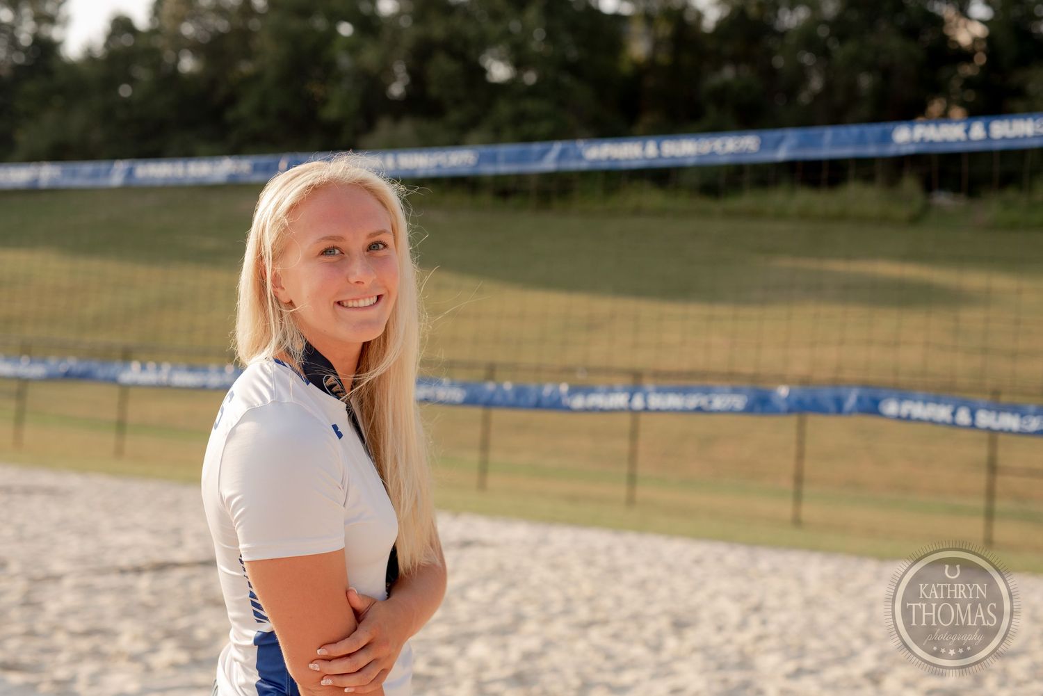 A volleyball player poses on a sandy court during a sunny day with athletic gear.