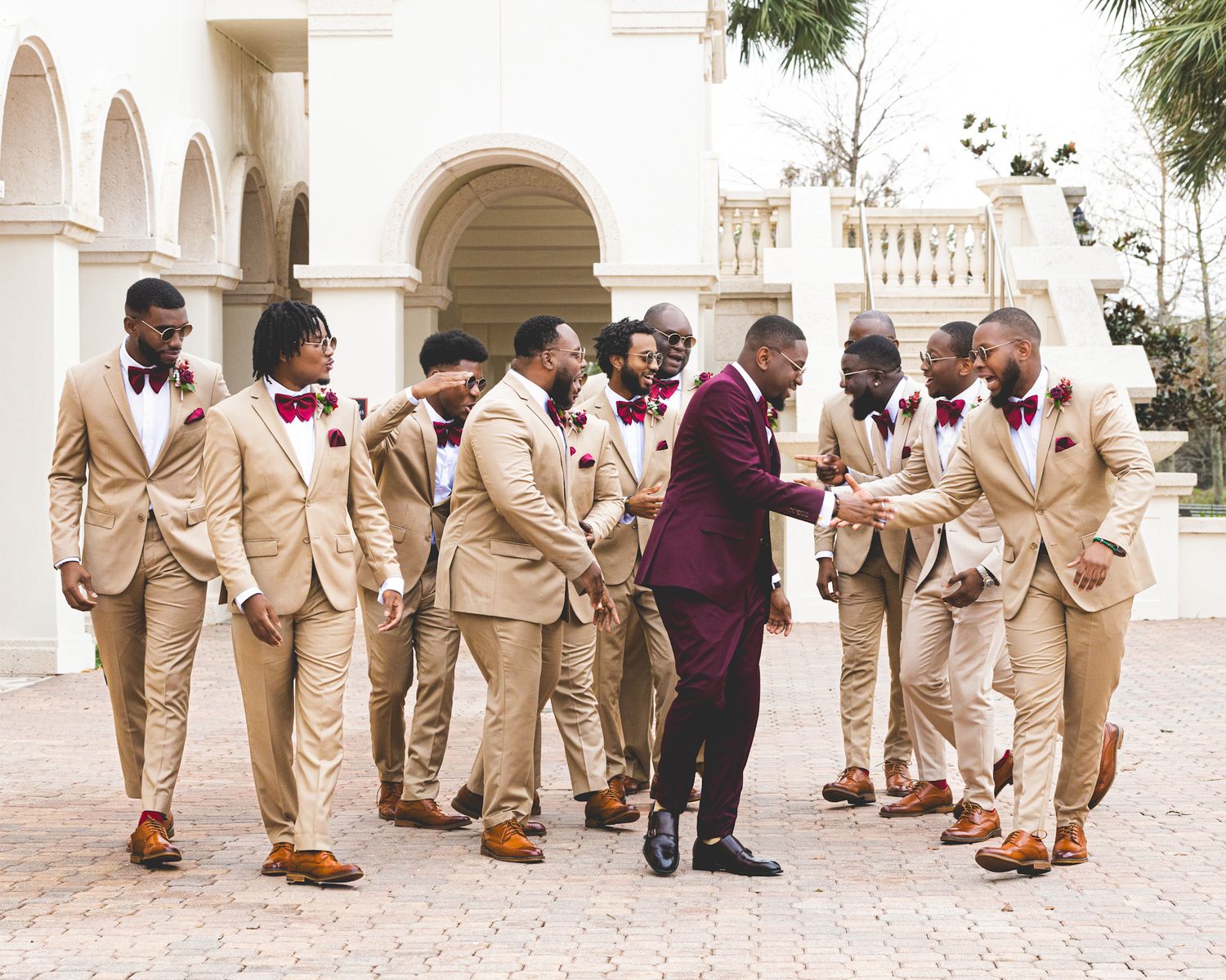 Wedding party in tan suits gathered around person in burgundy suit on stone courtyard.