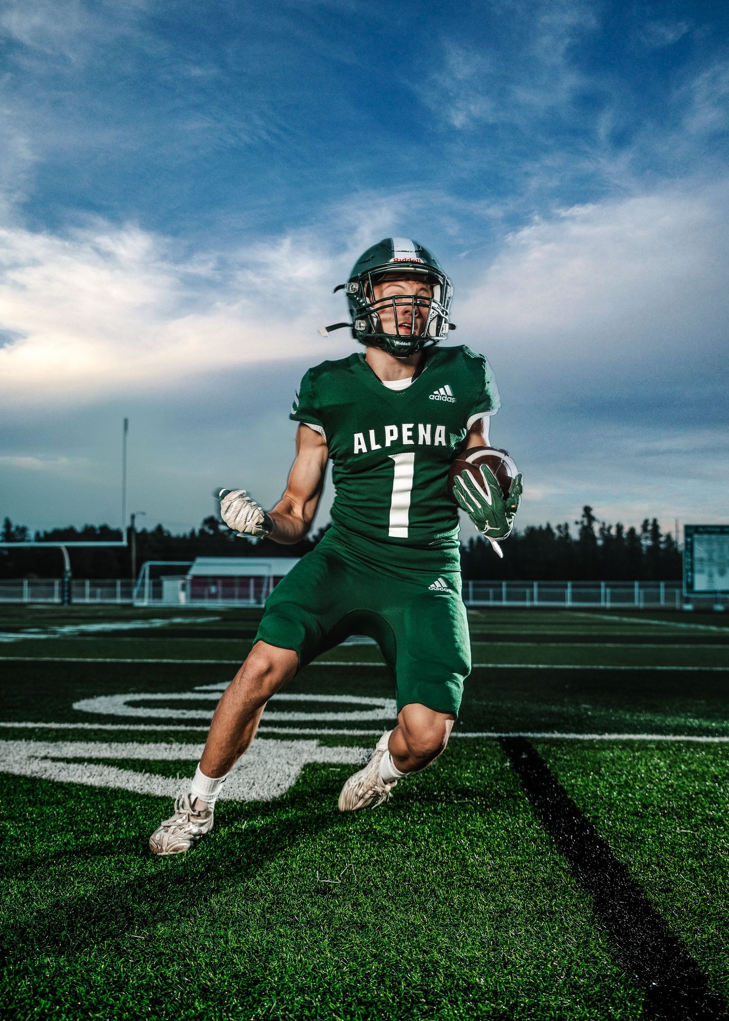 Action shot of high school football player in green Alpena uniform running on field under dramatic sky.