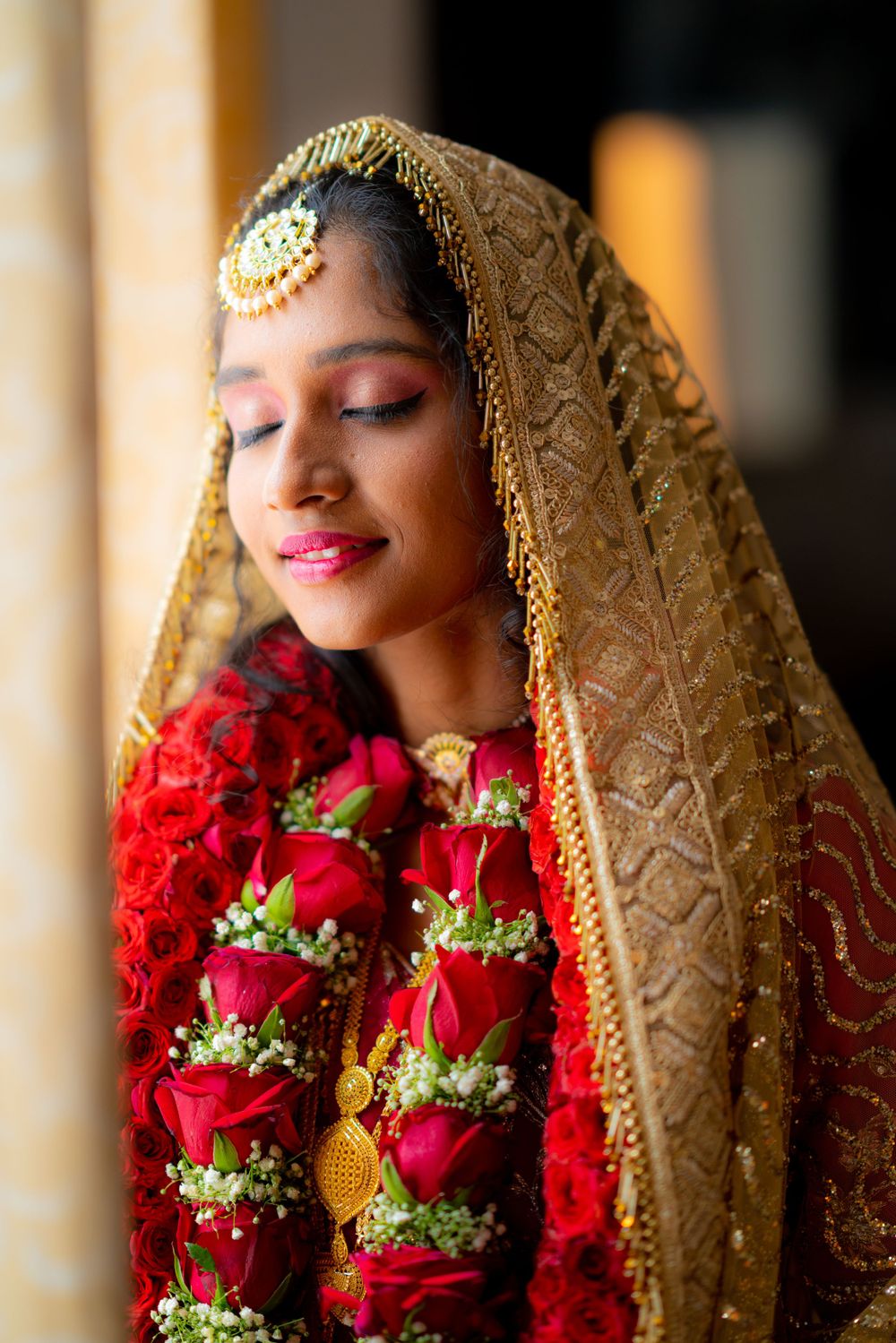 A bride wearing a golden veil and red rose garland glows in natural window light.
