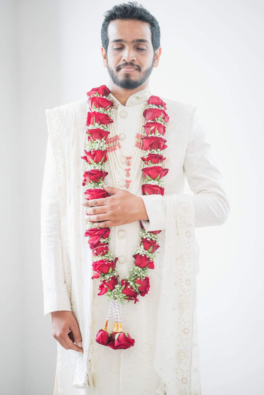 A groom wearing a white sherwani with a red floral garland poses against a light background.