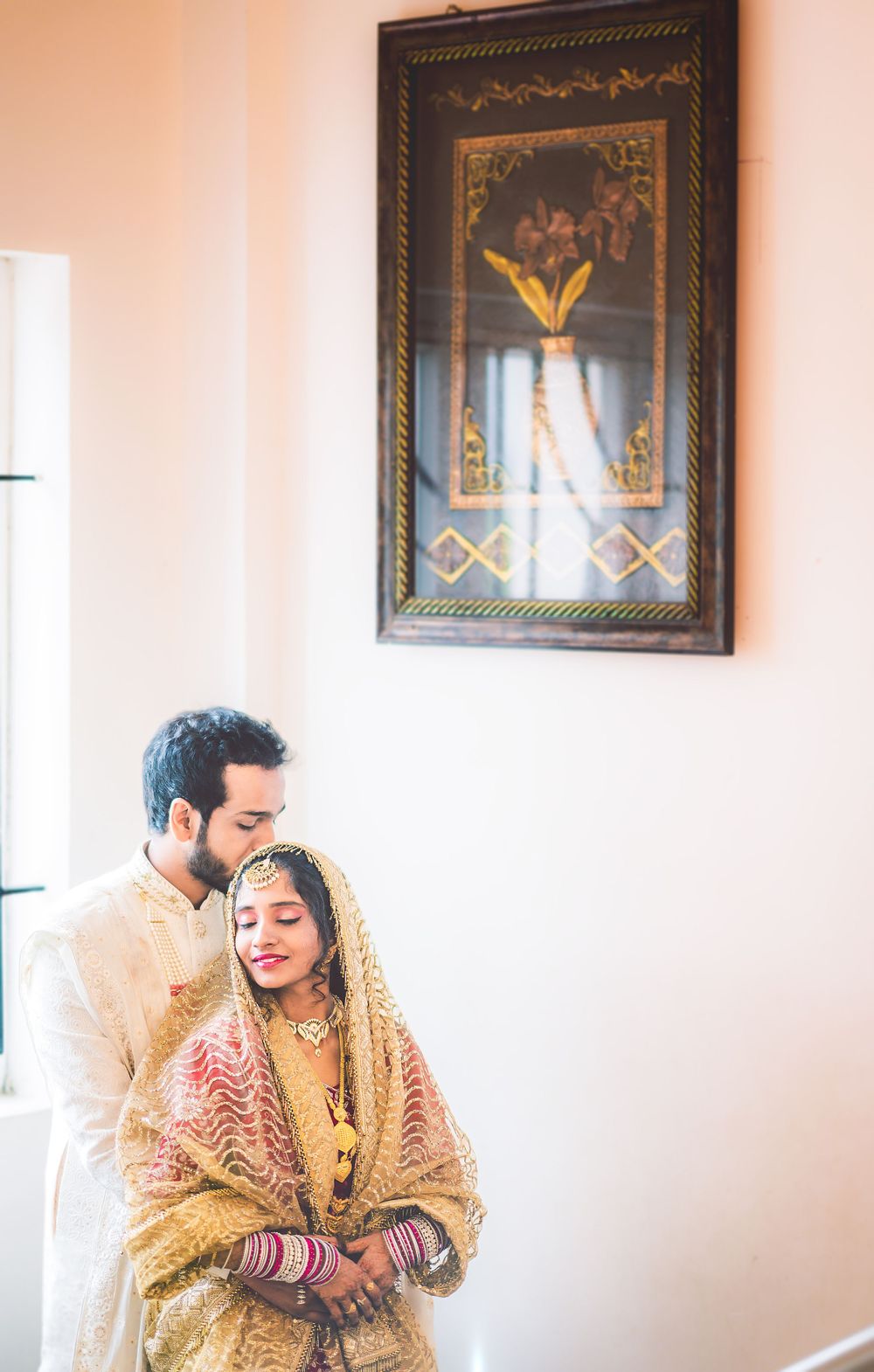 A romantic wedding couple shares an intimate moment under an ornate framed artwork.