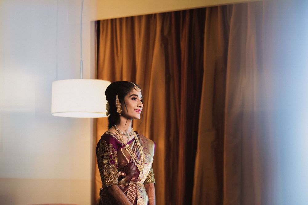 A traditional South Asian bride poses elegantly in wedding attire next to a table lamp with brown curtains in background.