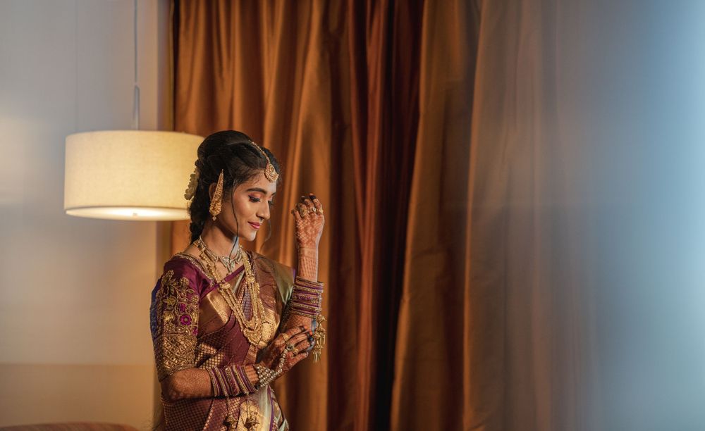 A bride in traditional South Asian attire poses gracefully beside a warm-lit lamp and brown curtains.