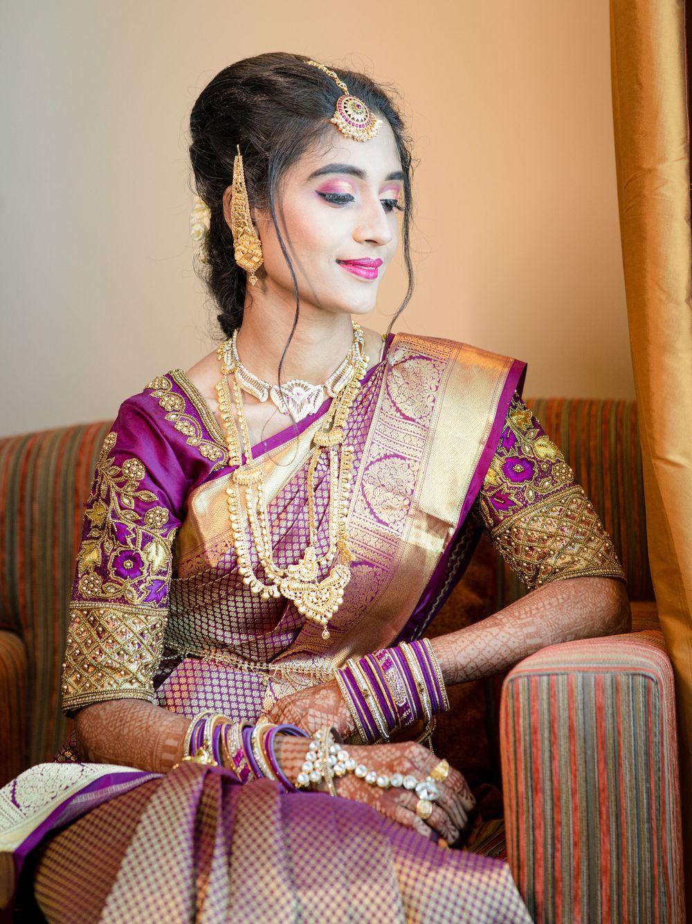 South Asian bride wearing purple and gold silk saree with traditional jewelry sits elegantly on a patterned couch.