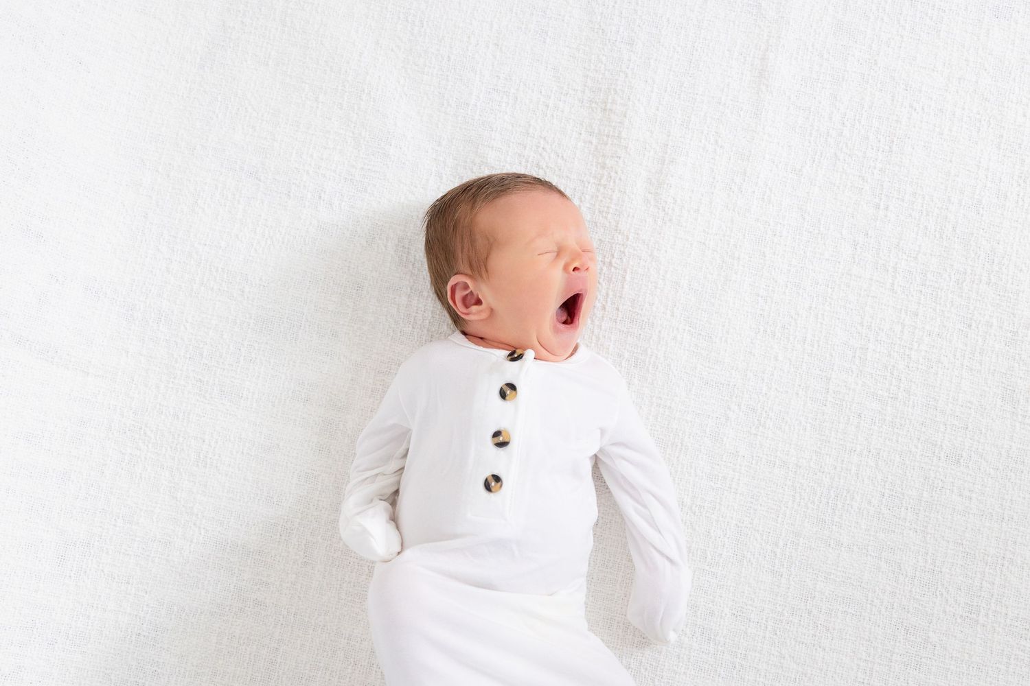 Newborn baby dressed in a white sleeper yawns.