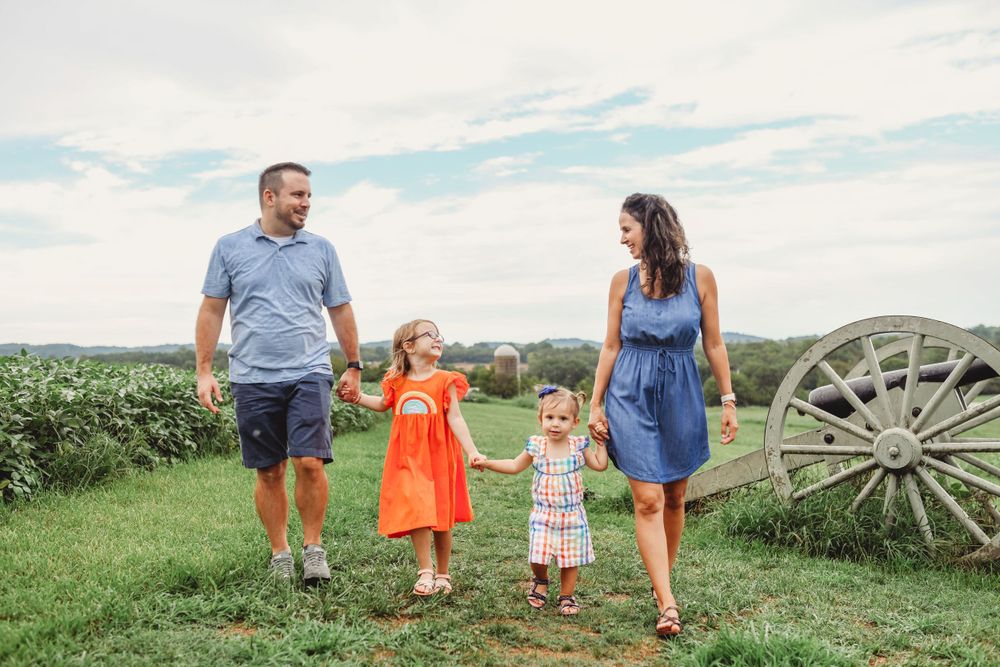 A family walks together on a grassy hill with an antique wagon wheel in the background on a sunny day.