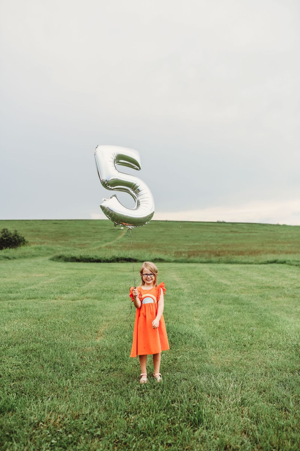 Child in orange dress playing with silver balloon letter in green field.