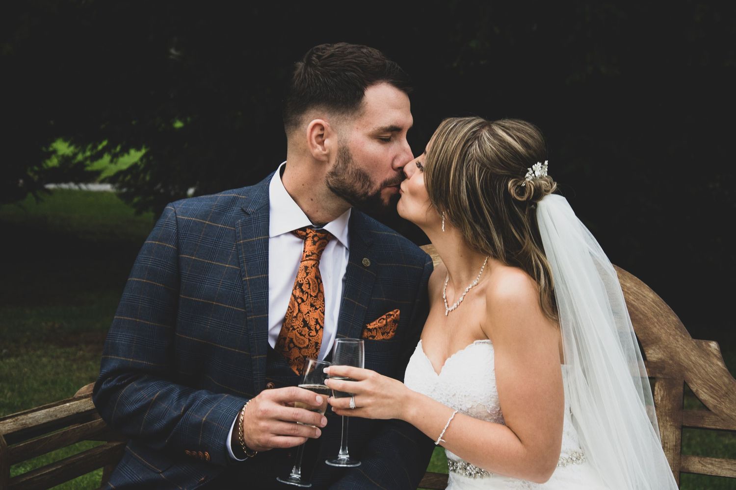 Newlyweds share a toast outdoors, bride in white dress with veil and groom in navy suit.