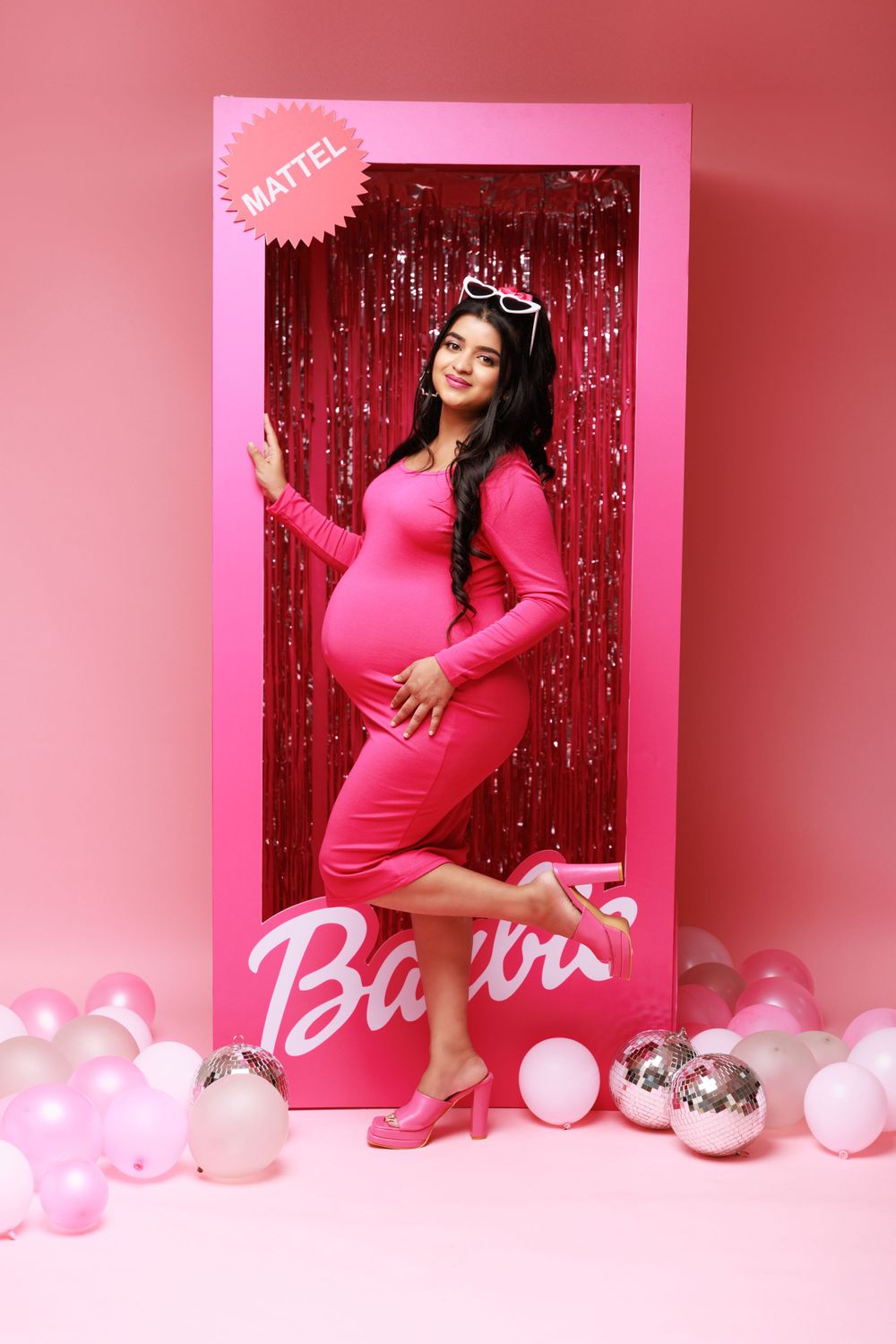 Creative maternity photo recreating Barbie doll box with pink dress and sparkly backdrop.