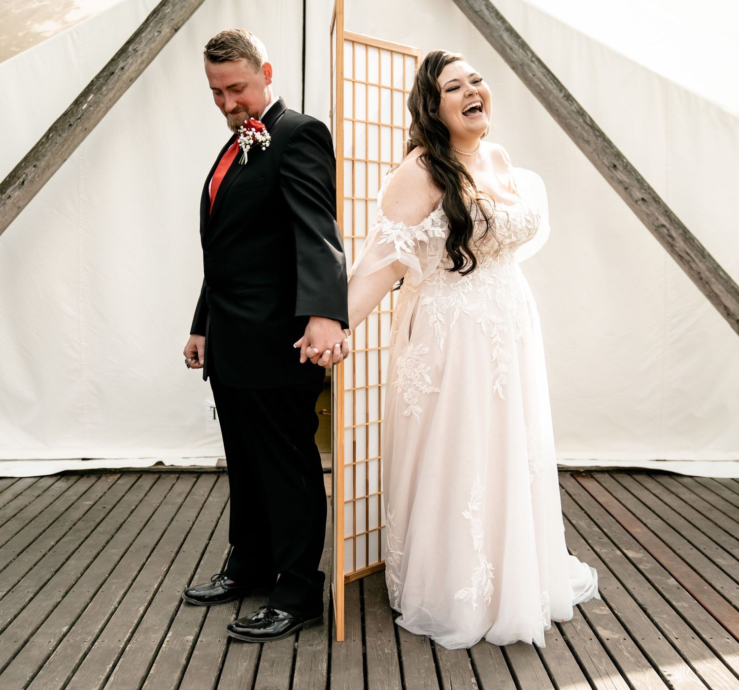 Couple in formal wedding attire standing back-to-back against white tent wall.