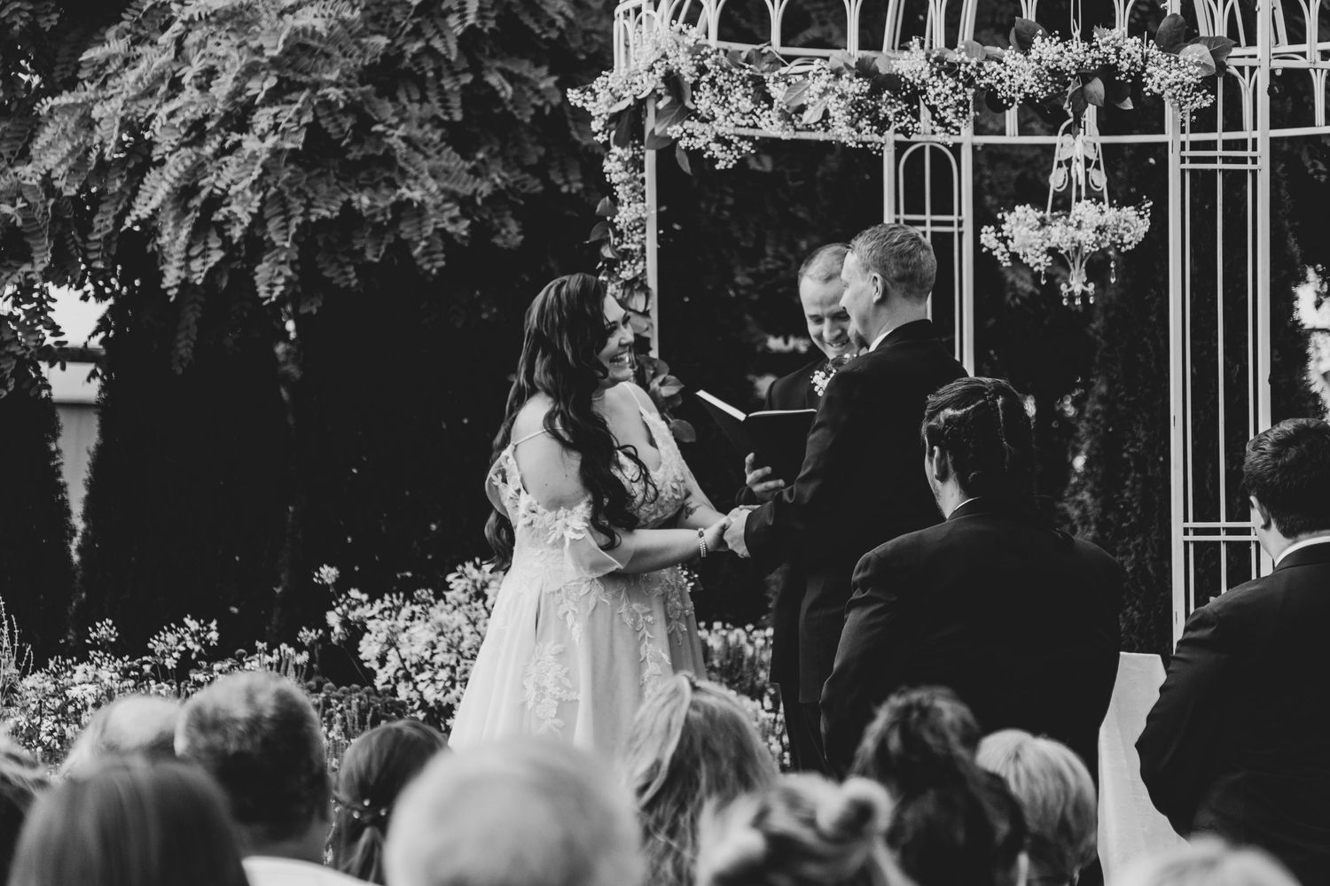 Black and white photo of outdoor wedding ceremony under floral archway.