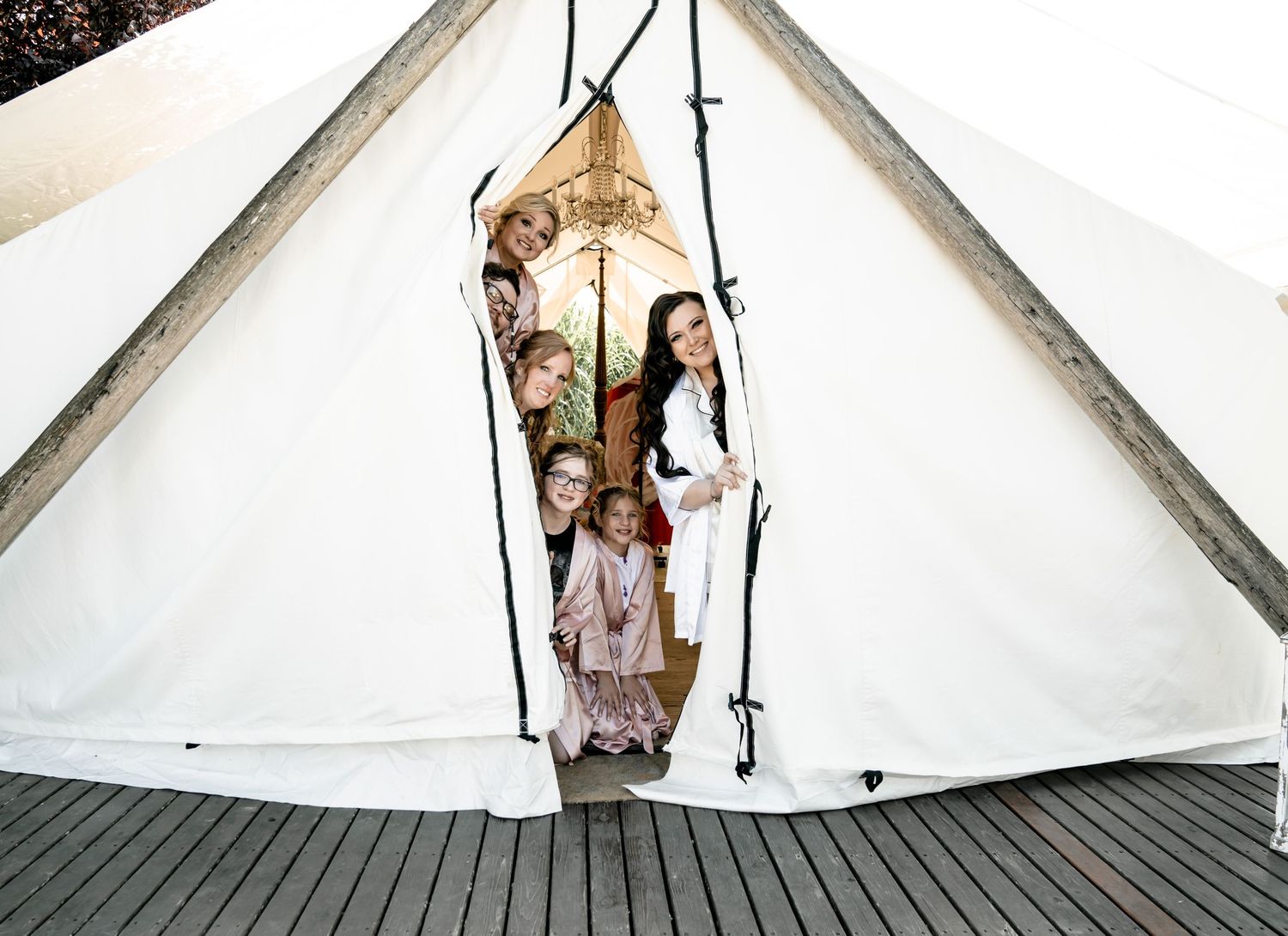 Wedding party peeking through entrance of large white tent on wooden deck.