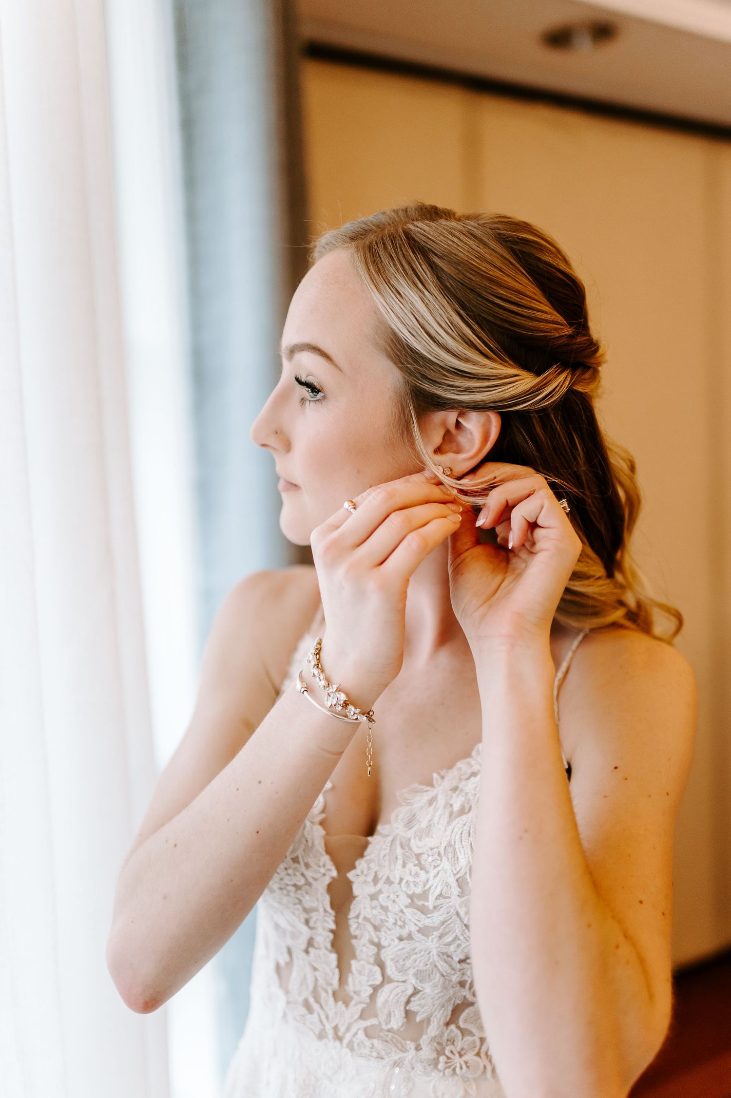 Bride adjusts jewelry while wearing an elegant lace wedding gown in natural window lighting.