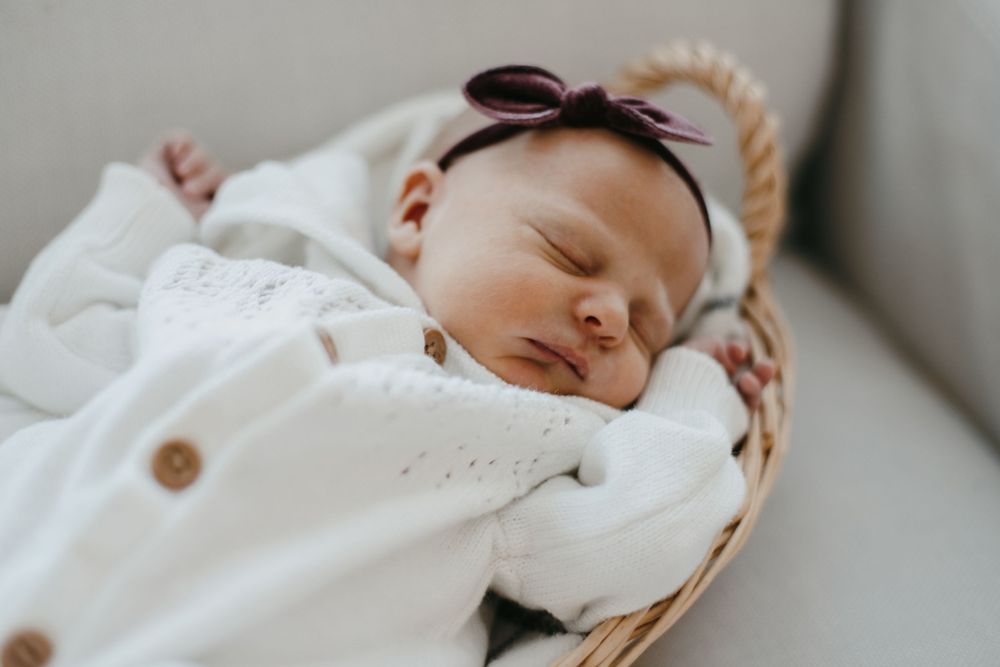 A sleeping baby wearing a white outfit and bow headband rests peacefully in a woven basket.