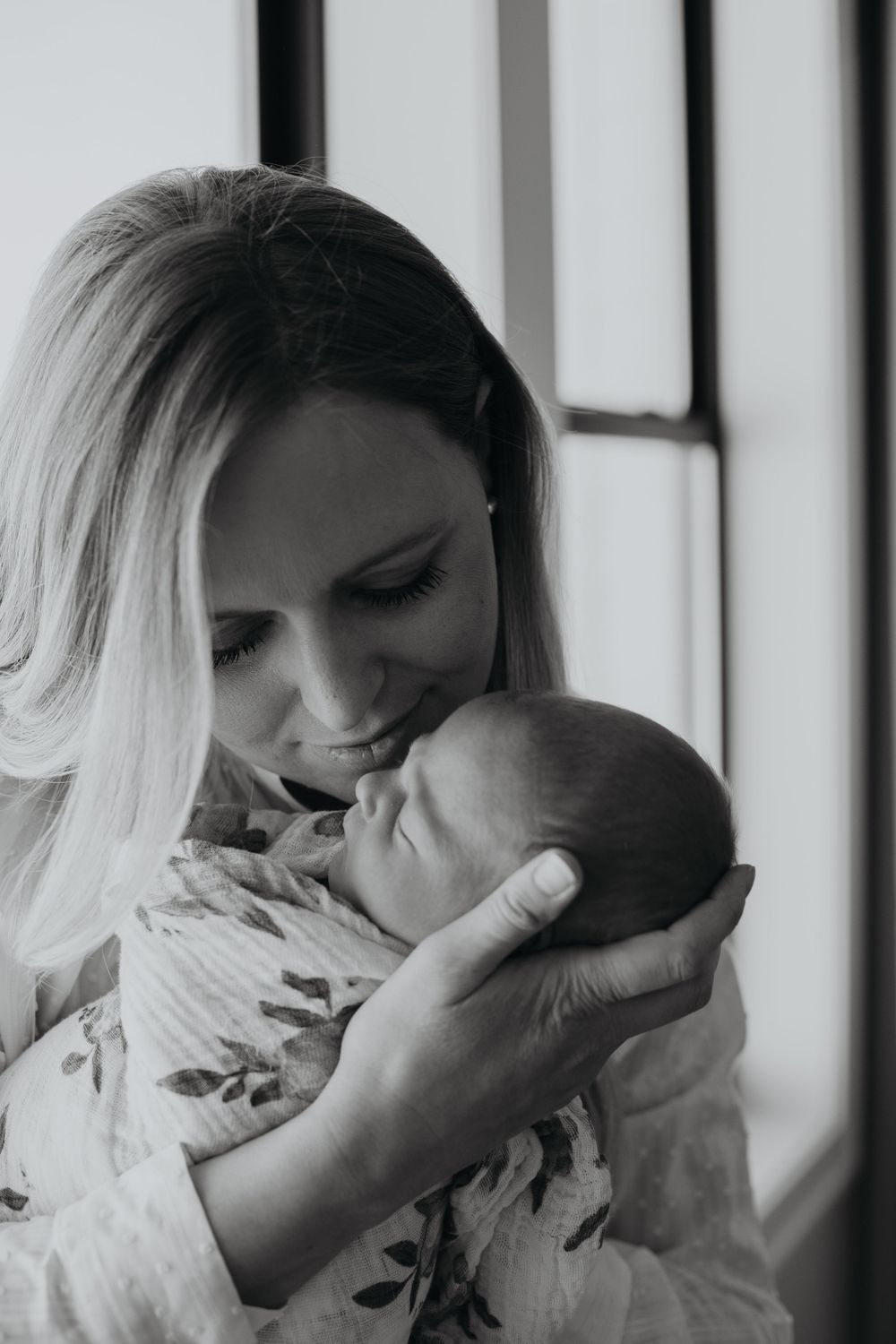 A tender black and white portrait of a mother holding her newborn baby near a window.