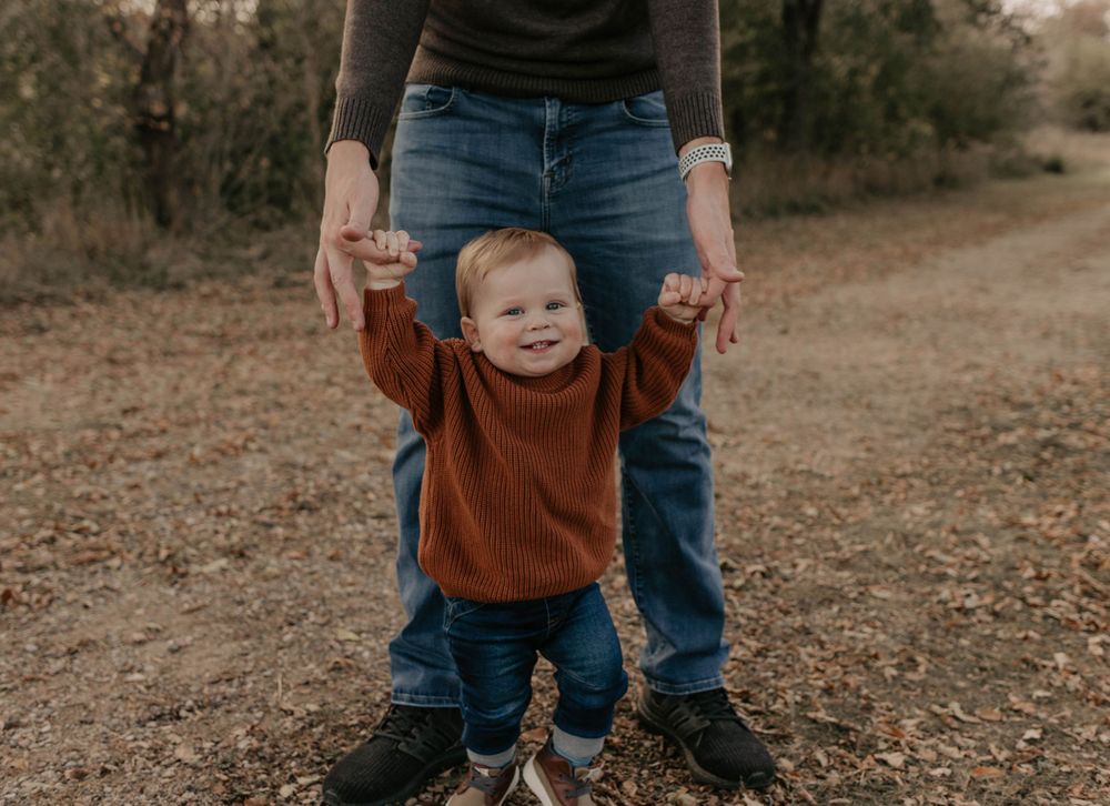 A toddler in a brown sweater learns to walk while holding an adult's hands on an autumn path.
