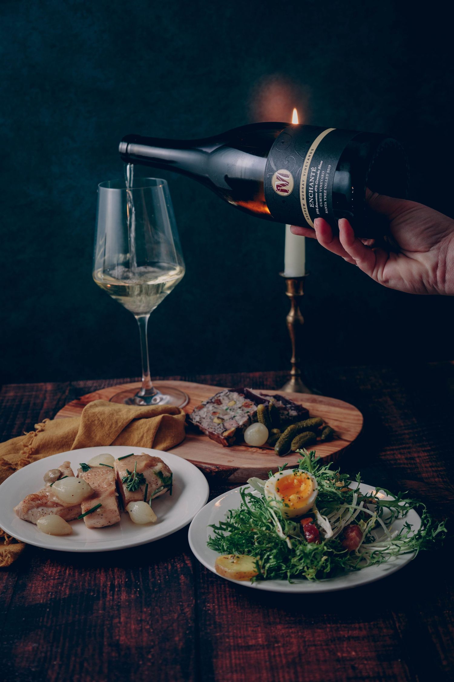 A hand pours white wine into a glass with food in the foreground