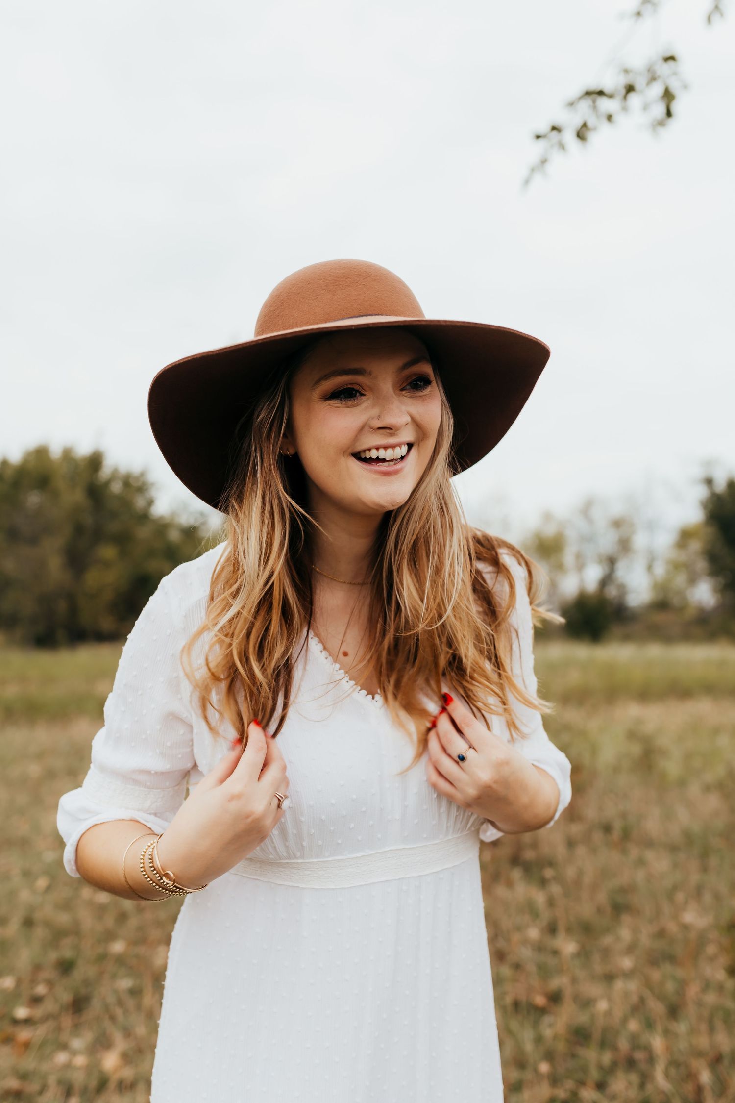 A person wearing a white dress and brown wide-brim hat smiles in an autumn field.