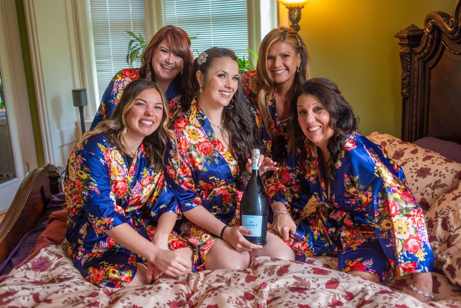 Bridesmaids in floral robes celebrating with champagne on bed during wedding morning.