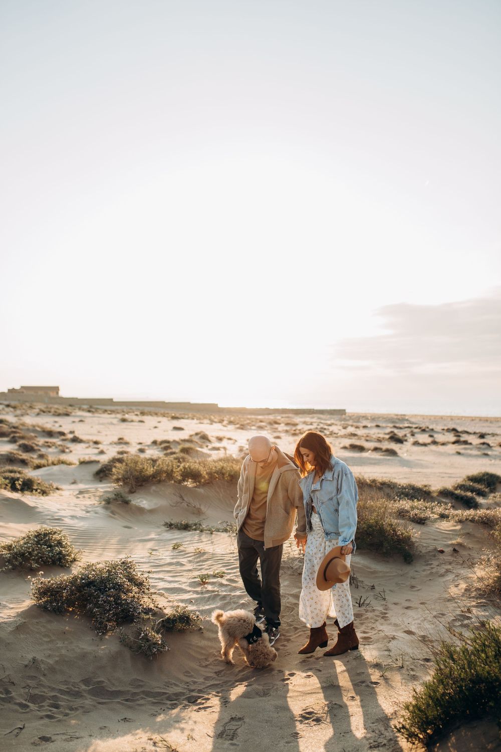 A couple strolls with their dog through desert dunes during golden hour.