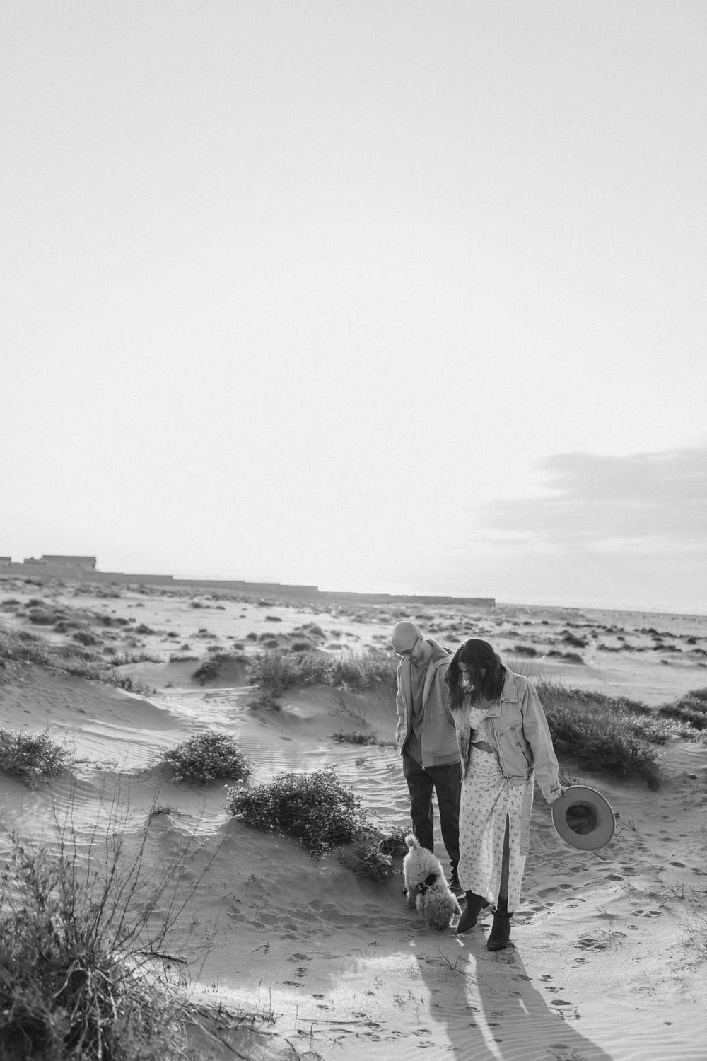 A couple walks with their small dog along sandy dunes in black and white.
