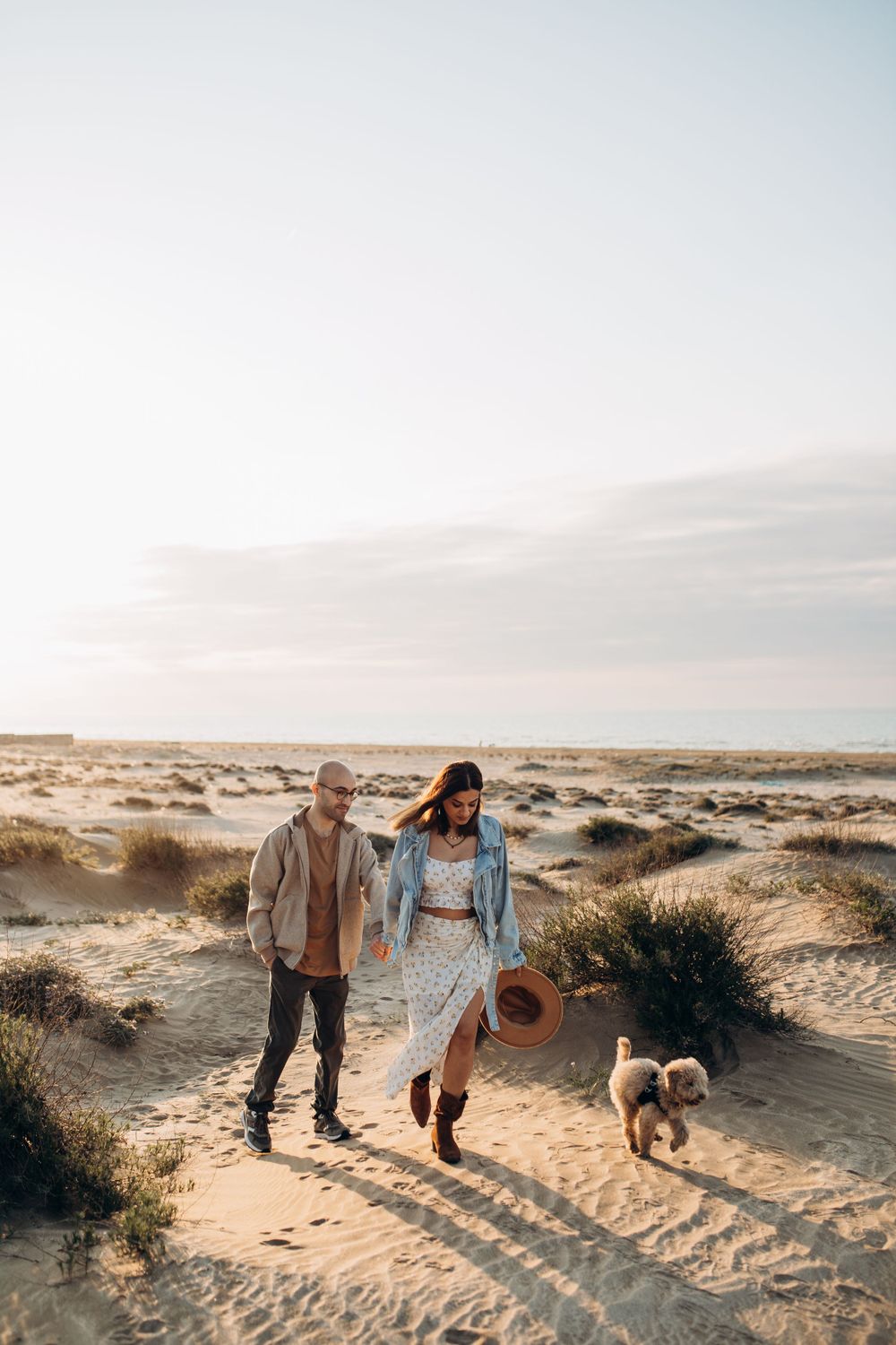 A couple in casual attire walking their dog through desert dunes at sunset.