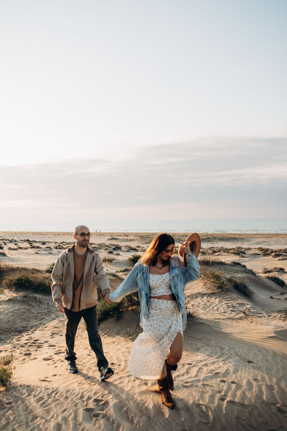 A couple walks hand in hand through sandy desert terrain at sunset.