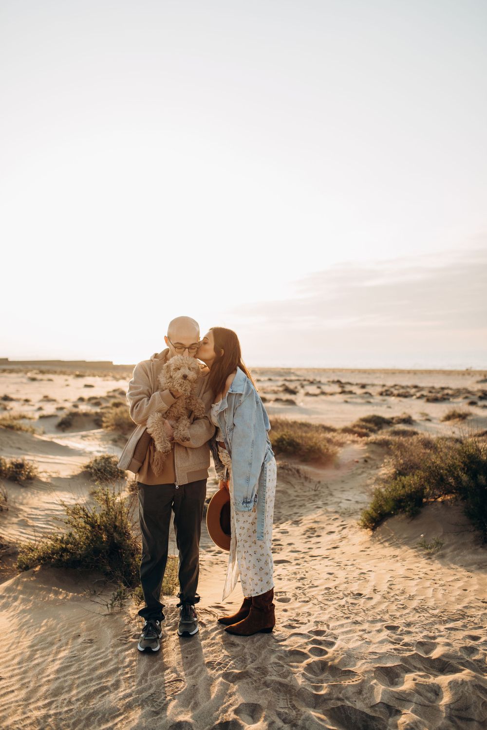A couple shares a tender moment with their fluffy dog while standing on sandy dunes at sunset.