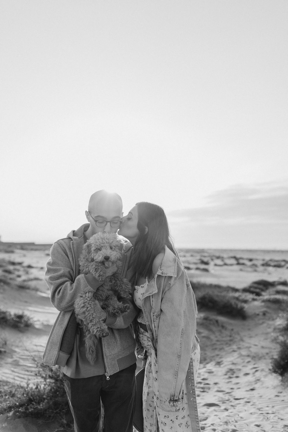 A couple holds their small curly-haired dog between them on a desert beach in black and white.