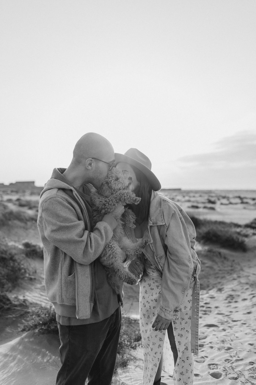 A romantic black and white photo of a couple cuddling their small dog in a desert landscape.