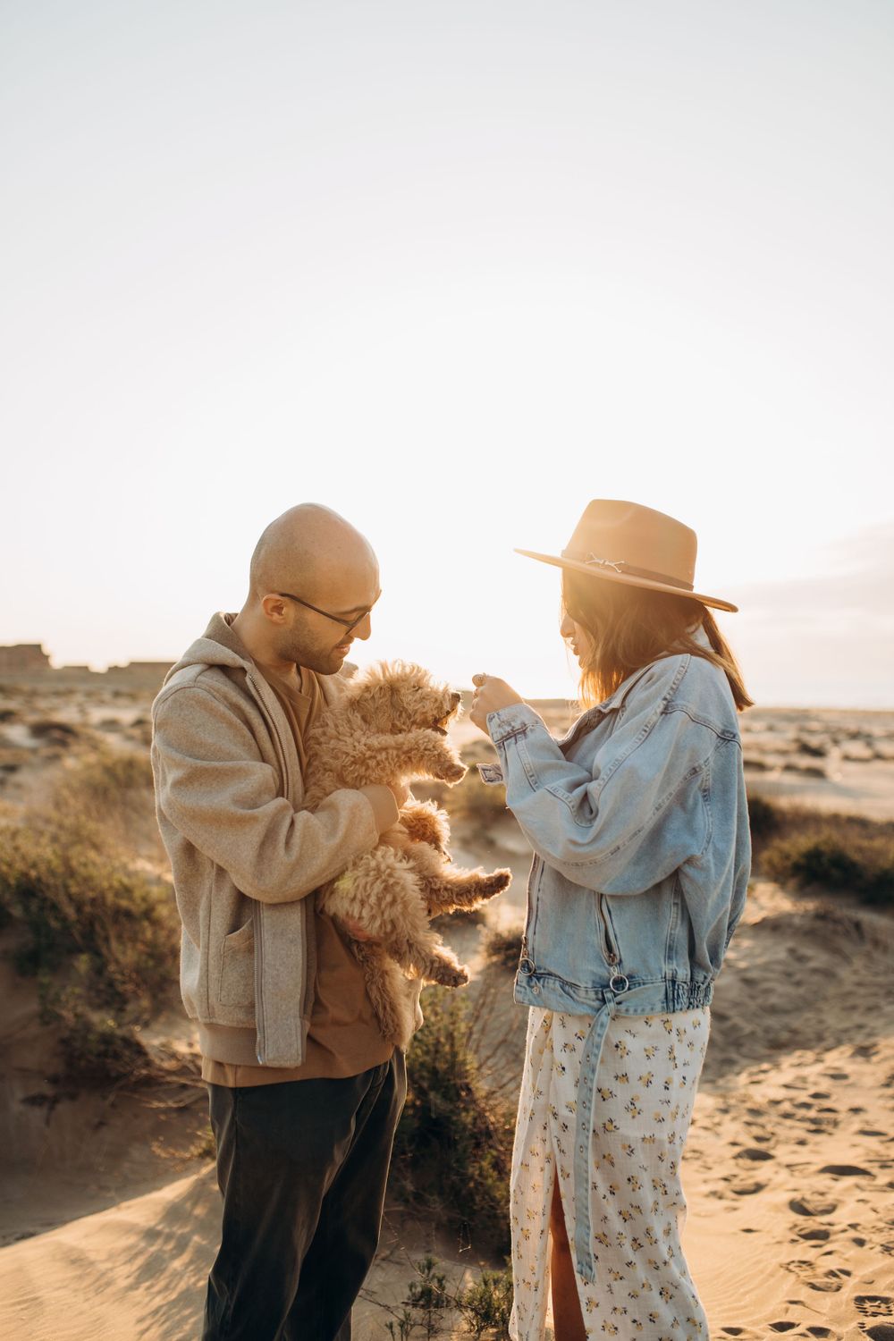 A couple plays with their small curly-haired dog against a desert backdrop at golden hour.