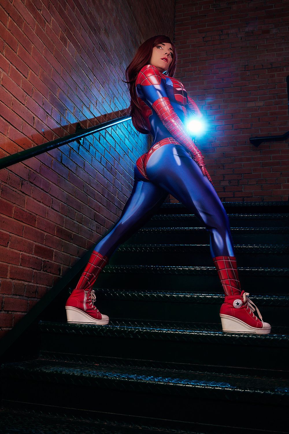 Woman in Spider-Man cosplay suit poses on dark staircase with dramatic blue and red lighting.