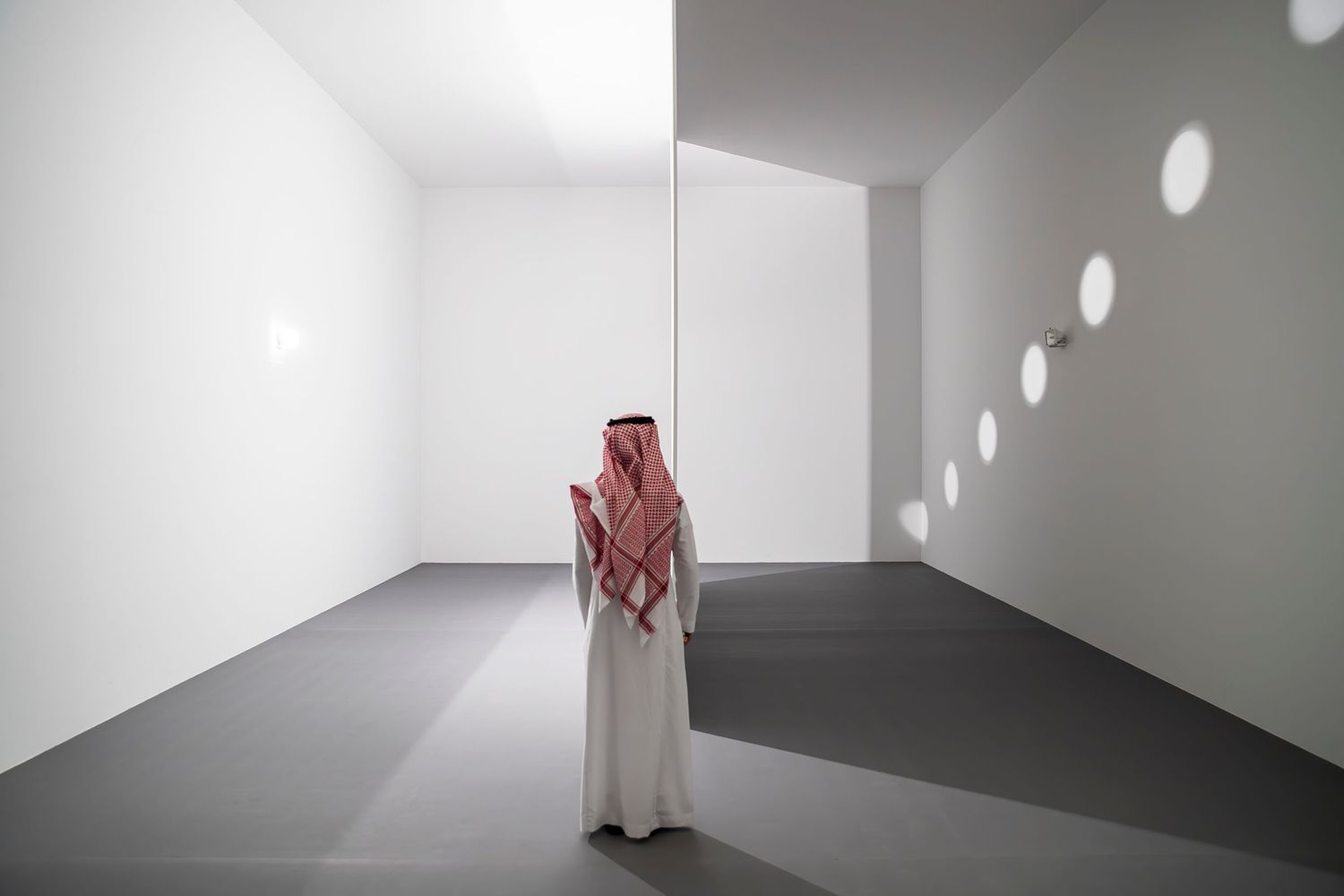 Figure stands in stark white gallery space with circular light projections on the wall.