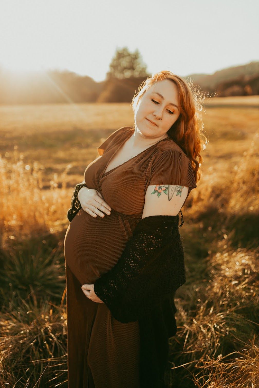 Pregnant redhead woman in brown dress cradling belly in golden autumn field.
