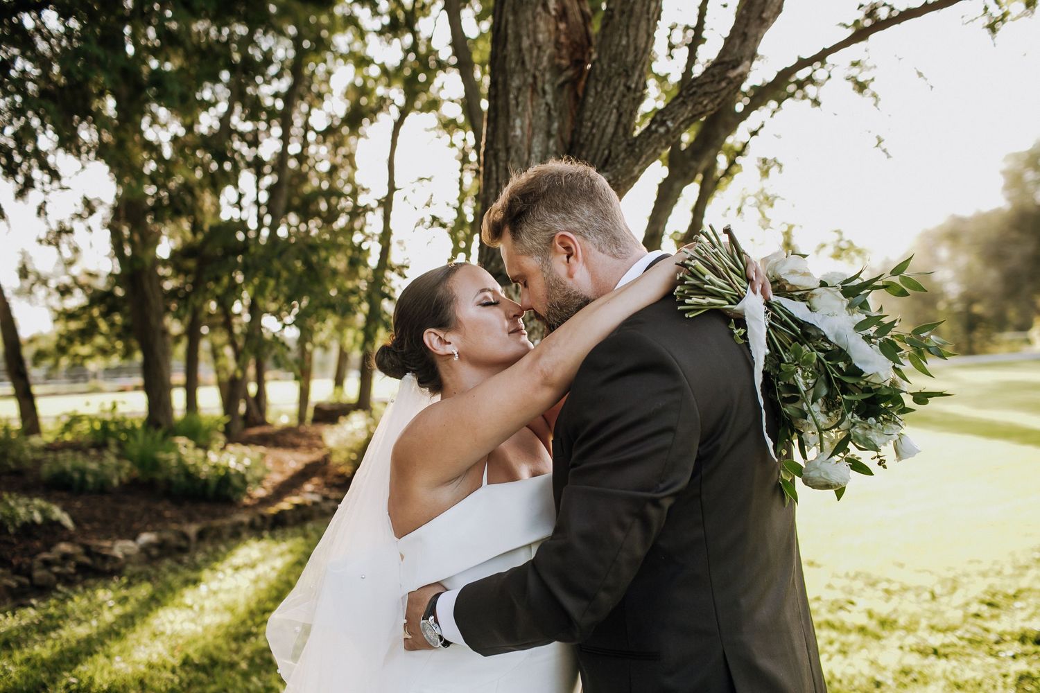 Newlywed couple sharing passionate kiss under trees in sunlit garden setting.
