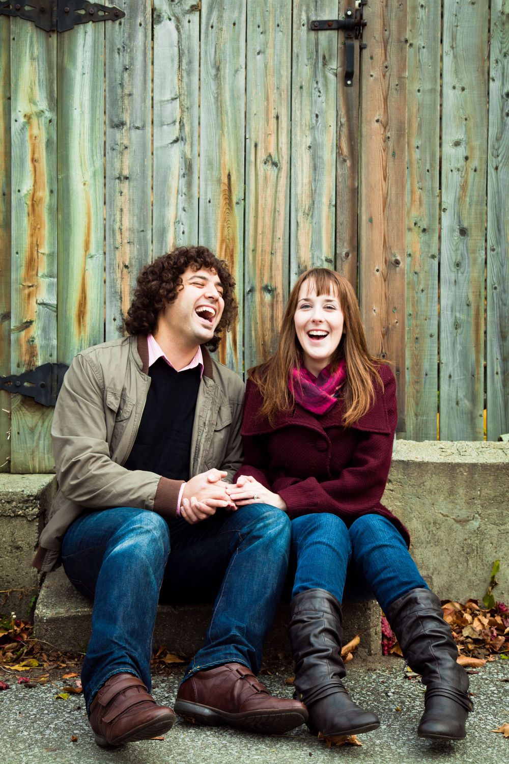 Couple laughing together while sitting against a weathered green wooden wall during their engagement session.