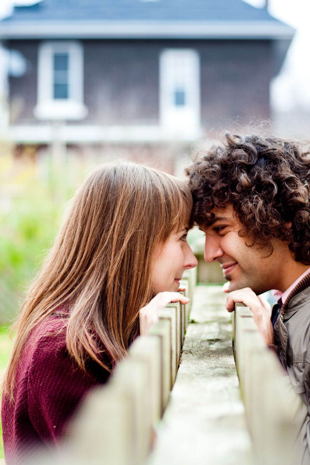 Couple sharing a close moment across a wooden fence with a house in the background during their engagement session.