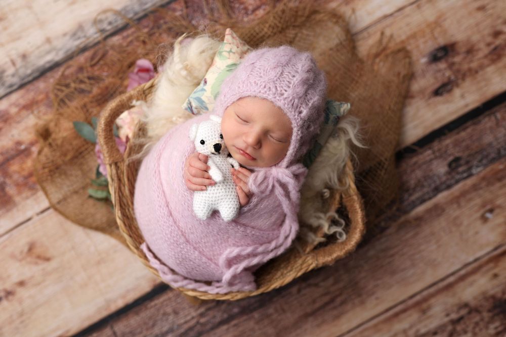 Newborn in pink swaddle and hat sleeping in basket with white plush toy.
