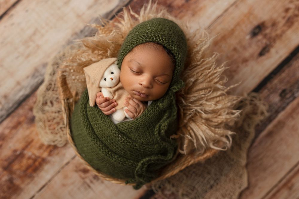 Sleeping newborn in green knit wrap with white teddy bear on textured backdrop.
