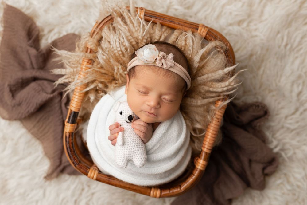 Sleeping newborn swaddled in white with floral headband in wicker basket.