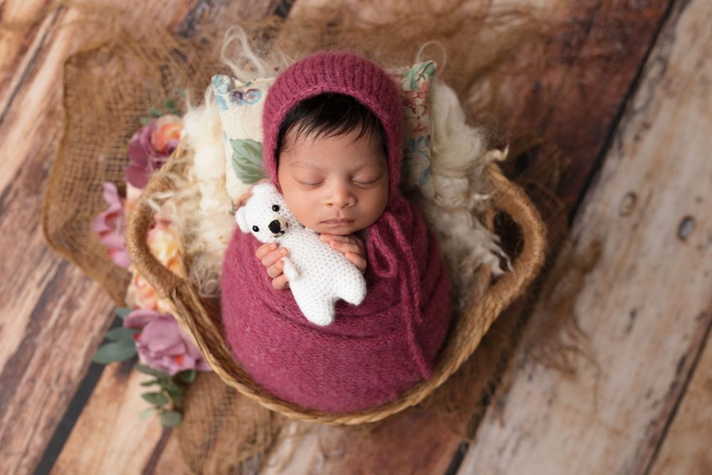 Sleeping newborn wrapped in burgundy swaddle with small teddy bear in rustic basket.