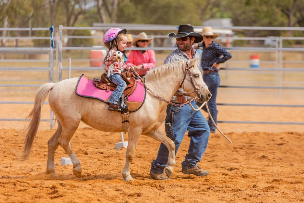 A rider in pink sits atop a light-colored pony while being led by an instructor in a dusty arena.