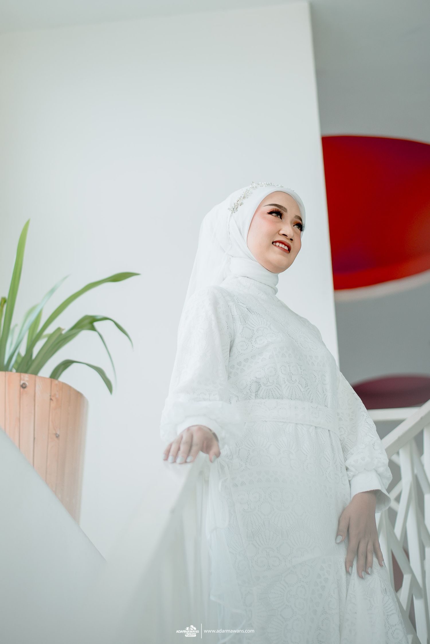 Elegant bride in white hijab and lace dress posing on modern staircase.